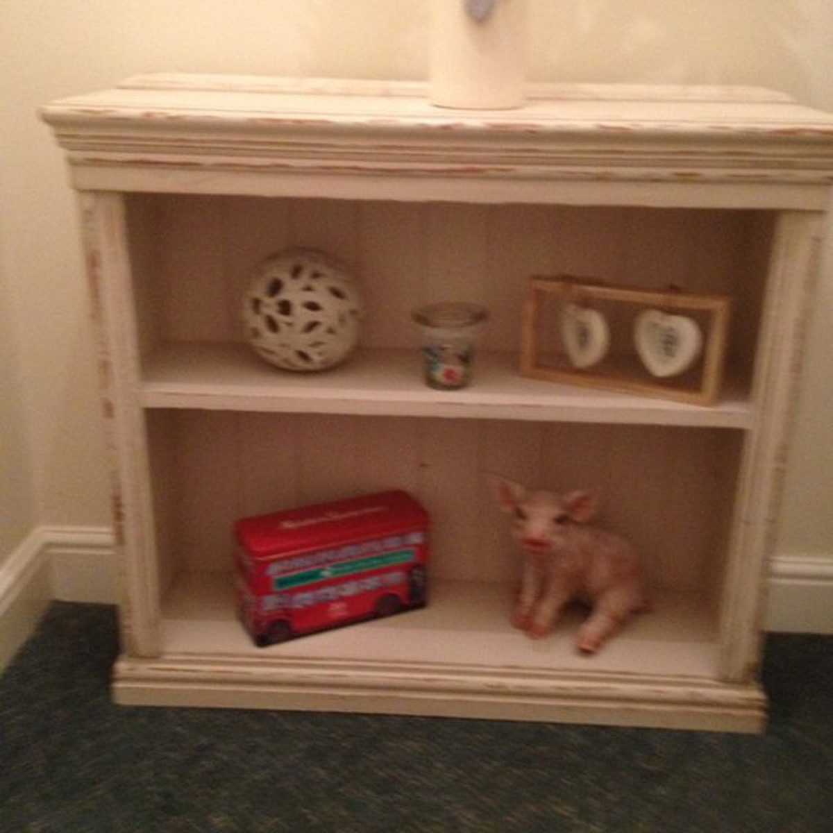 A white wooden shelf displays decorative items, including a ceramic sphere, candle, photo frames, a toy bus, and a small pig figurine, against a neutral wall.
