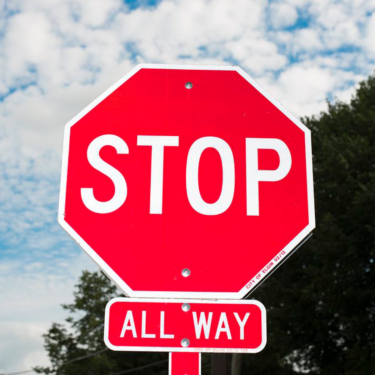 A red octagonal stop sign is prominently displayed, instructing drivers to halt. The background features a partly cloudy sky and green foliage.