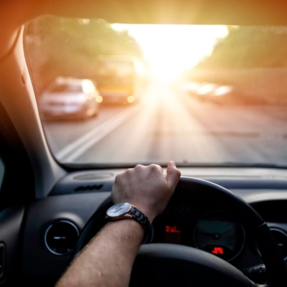 A hand grips the steering wheel while driving towards a sunlit road, surrounded by faint outlines of vehicles in the distance.