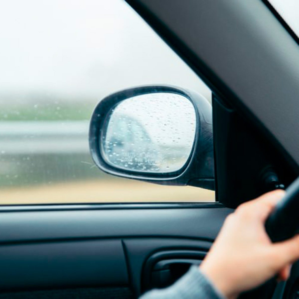 A hand grips the steering wheel while rain-covered side mirrors reflect a blurred road, indicating a vehicle in motion during a rainy day.