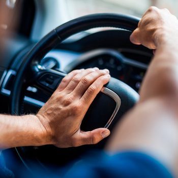 A hand is gripping a car steering wheel, while another hand rests on the wheel, indicating the driver is engaged in operating the vehicle.