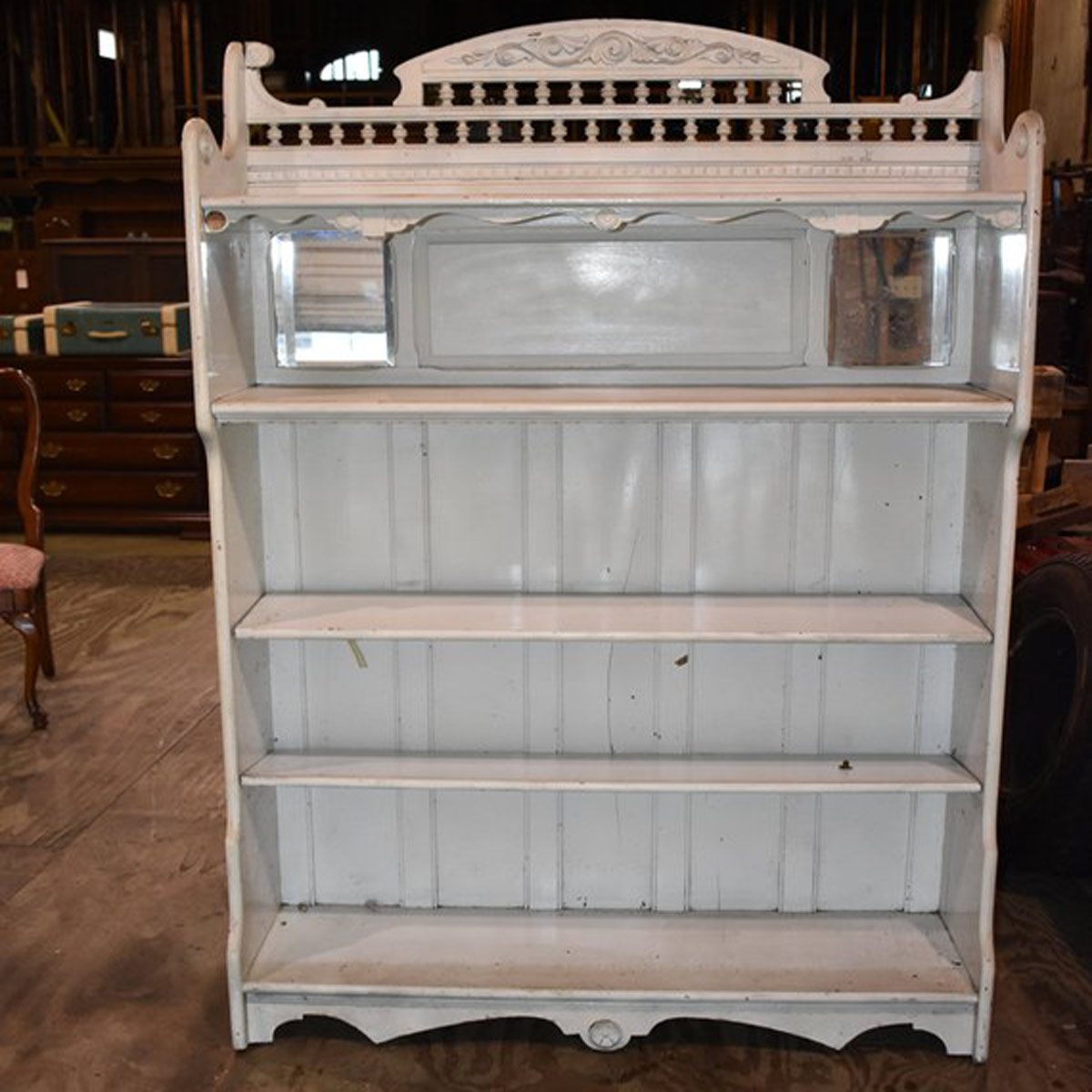 A white wooden shelf with multiple open shelves stands upright, reflecting light from a nearby source in a rustic, wooden environment.