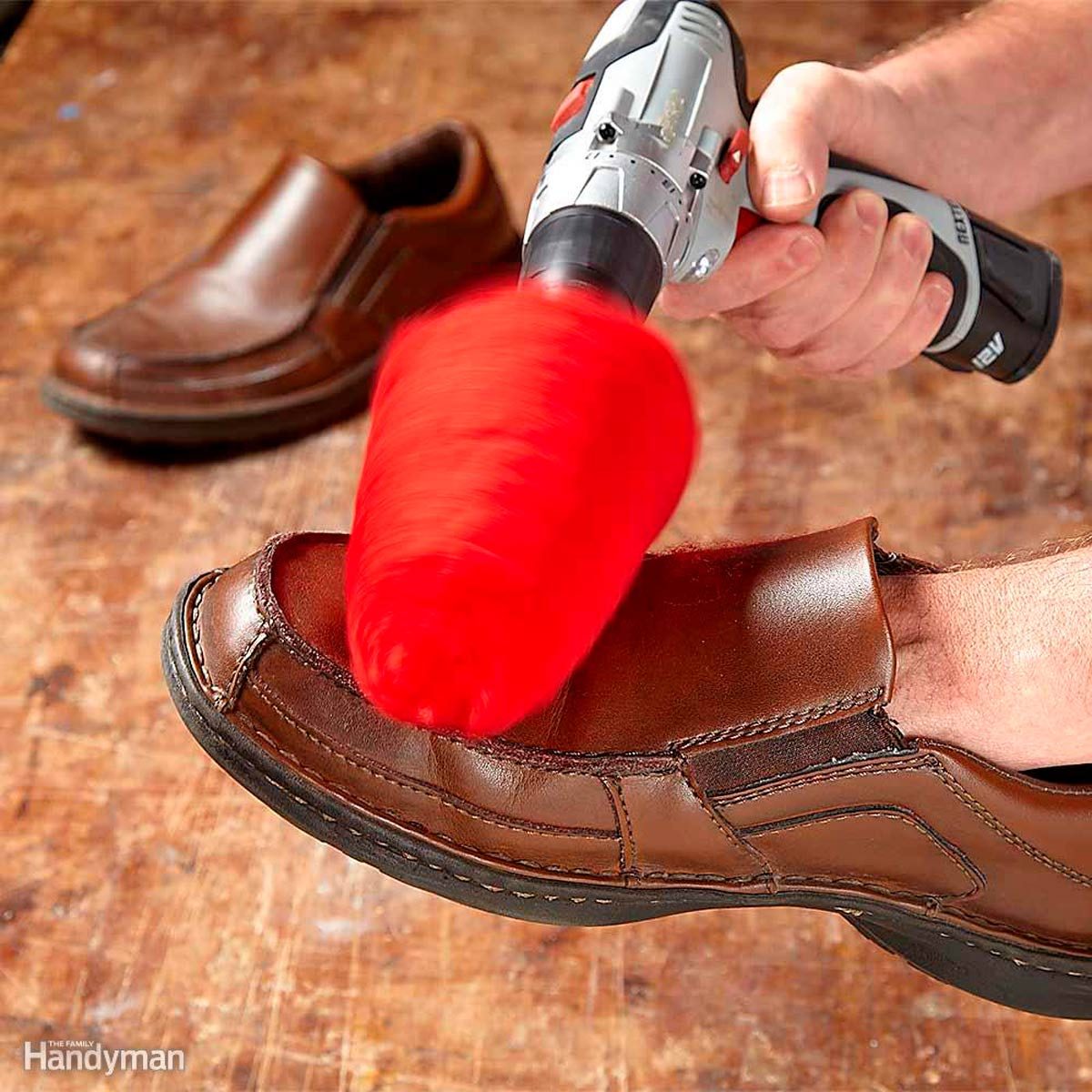 A hand-held cleaning tool polishes a brown shoe while a foot wears another shoe on a wooden surface. A second shoe is nearby.