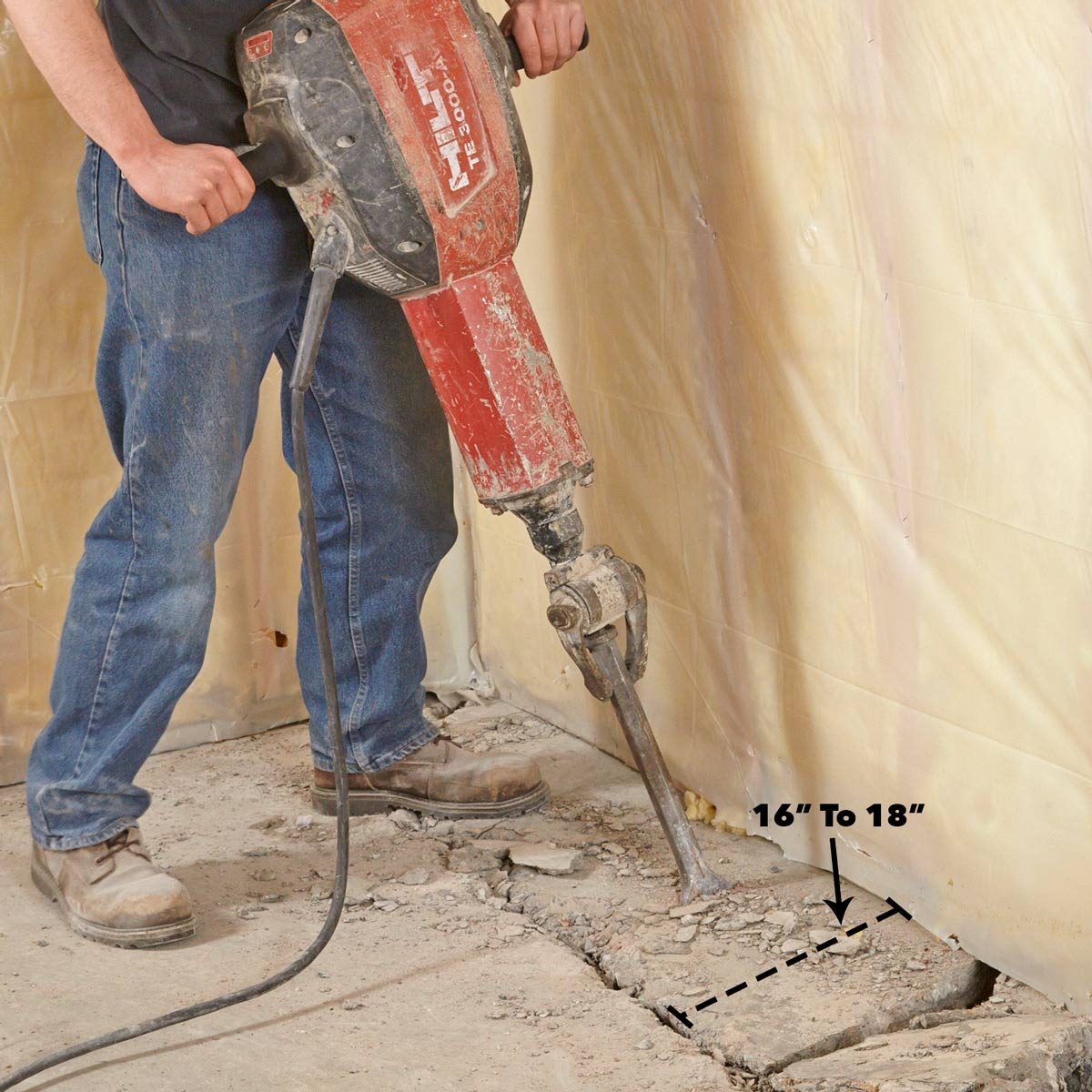 A person operates a jackhammer, breaking concrete along a wall. The environment appears to be a construction site with exposed flooring and temporary wall protection.