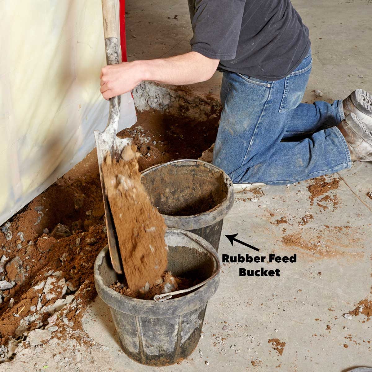 A person kneels, using a shovel to transfer dirt into two buckets on a concrete floor, surrounded by scattered debris and a plastic barrier.