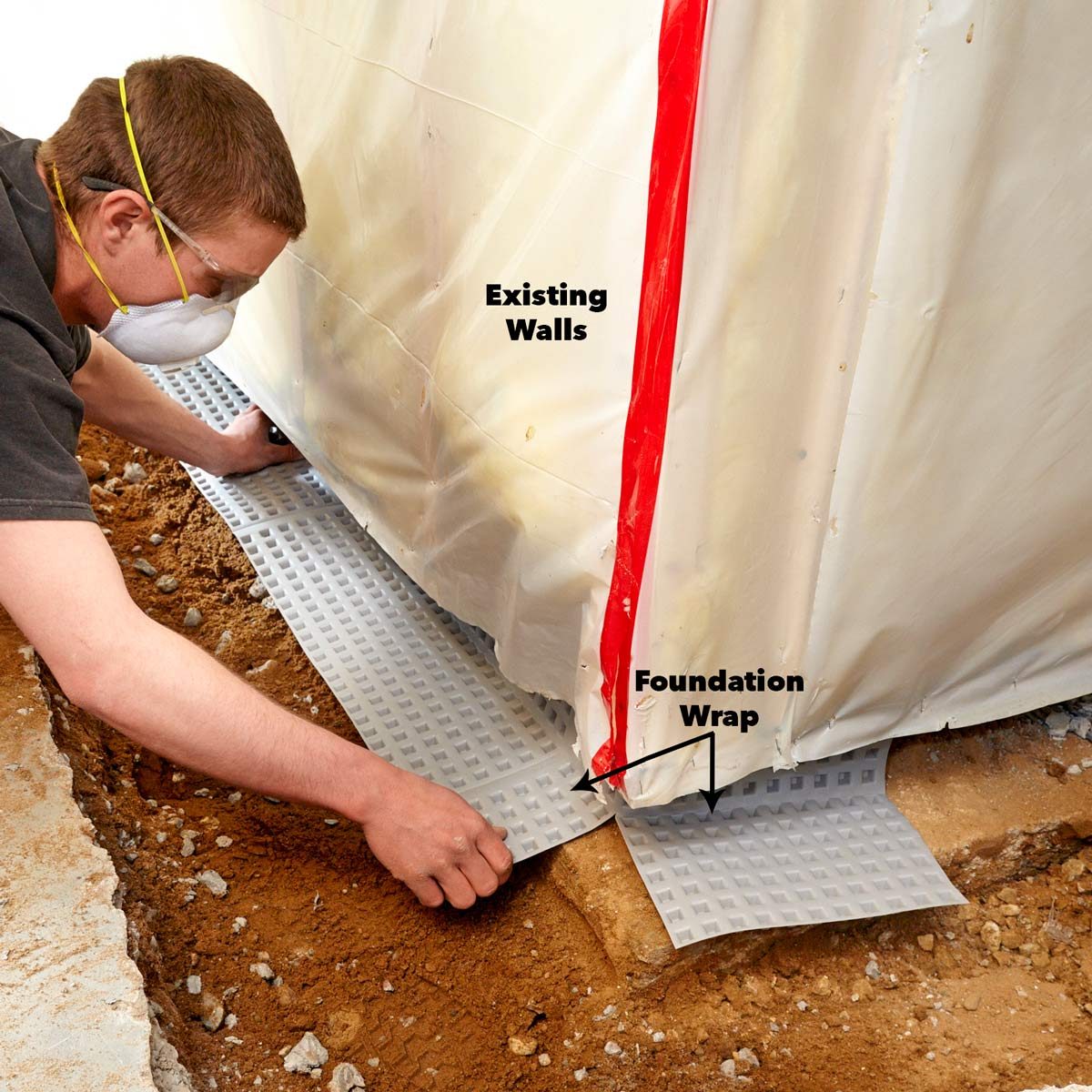 A person installs a foundation wrap along the base of existing walls in a dirt-filled environment, focusing on preventing moisture damage.