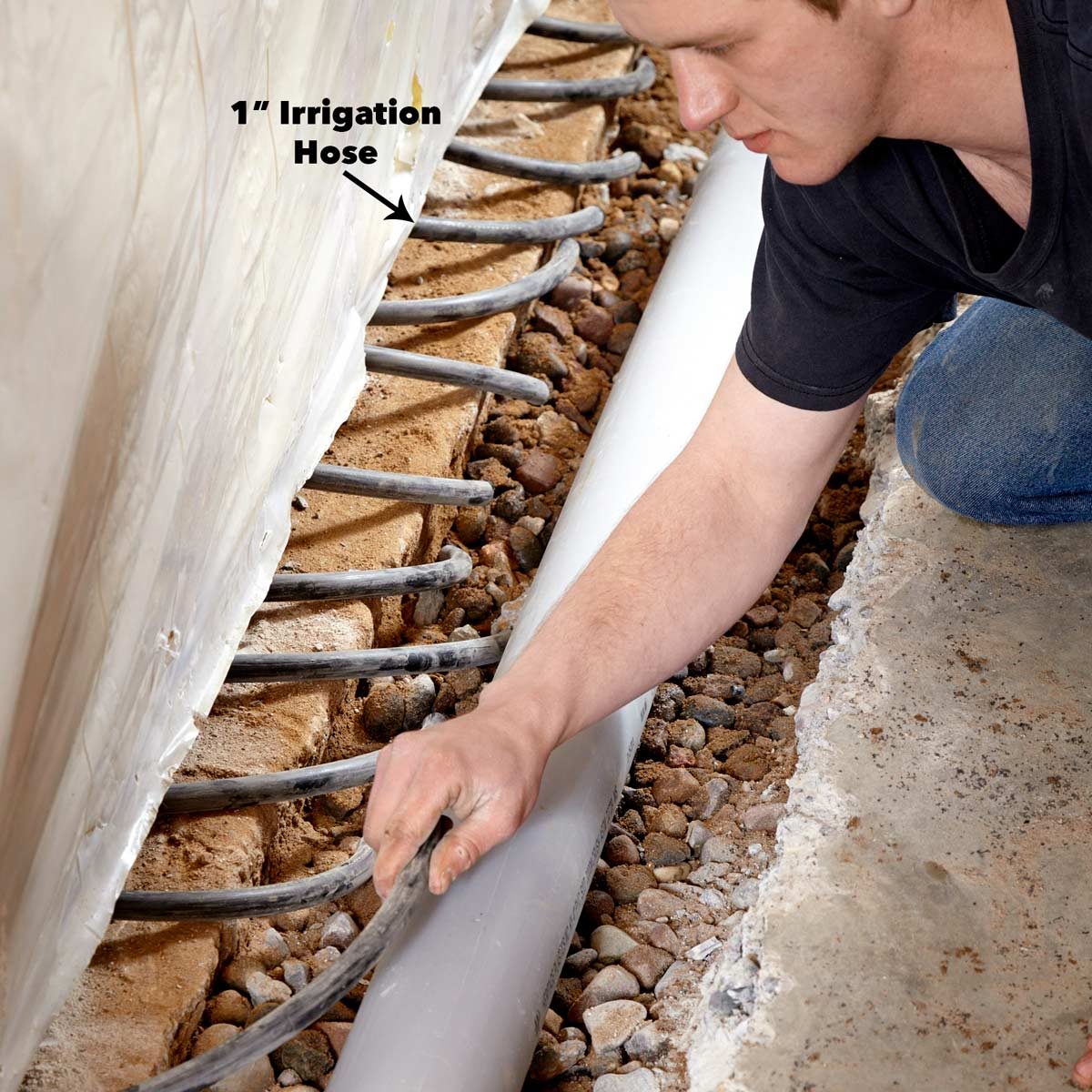 A person is adjusting a 1-inch irrigation hose near a rocky, uneven surface inside a construction area, using one hand for precision.