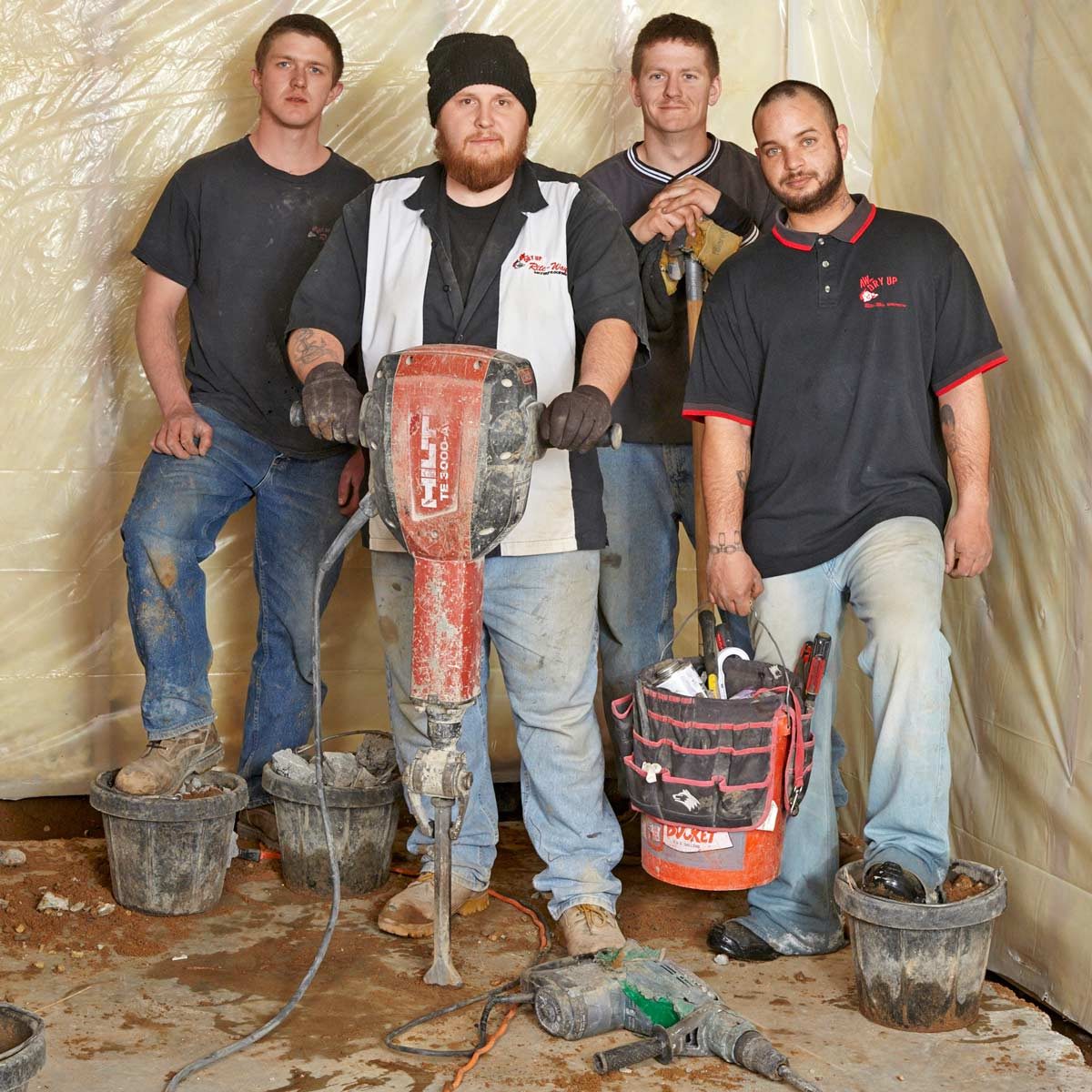 Four construction workers stand together, one holds a drill. They are in a partially covered work area with dirt and buckets around them.