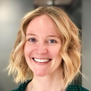 A smiling woman with wavy, blonde hair is facing the camera in a softly lit indoor space, wearing a dark green checkered top.