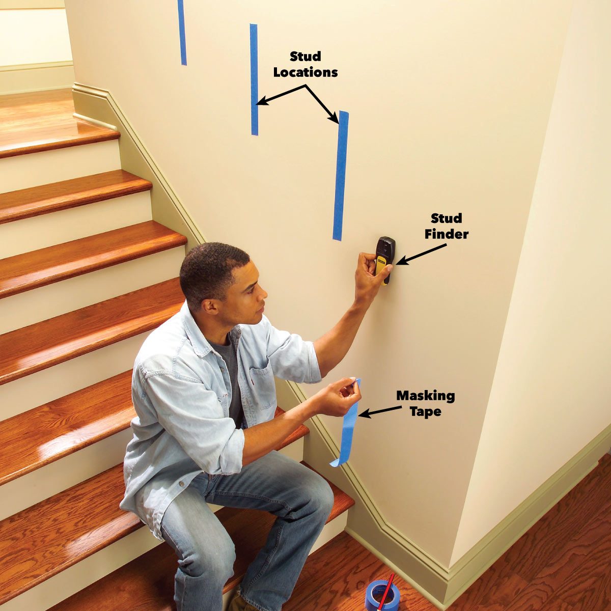 A man uses a stud finder on a wall while applying masking tape, with blue tape indicating stud locations in a staircase setting.