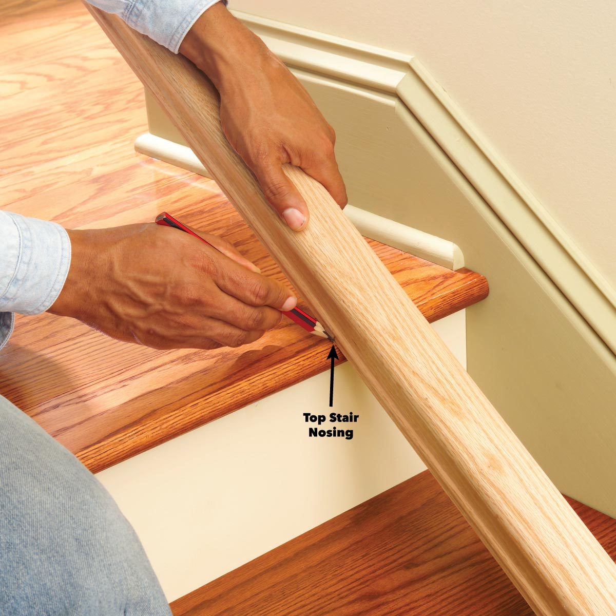 A person holds a wooden handrail while marking a measurement on the top stair nosing, surrounded by polished hardwood flooring and a light-colored wall.