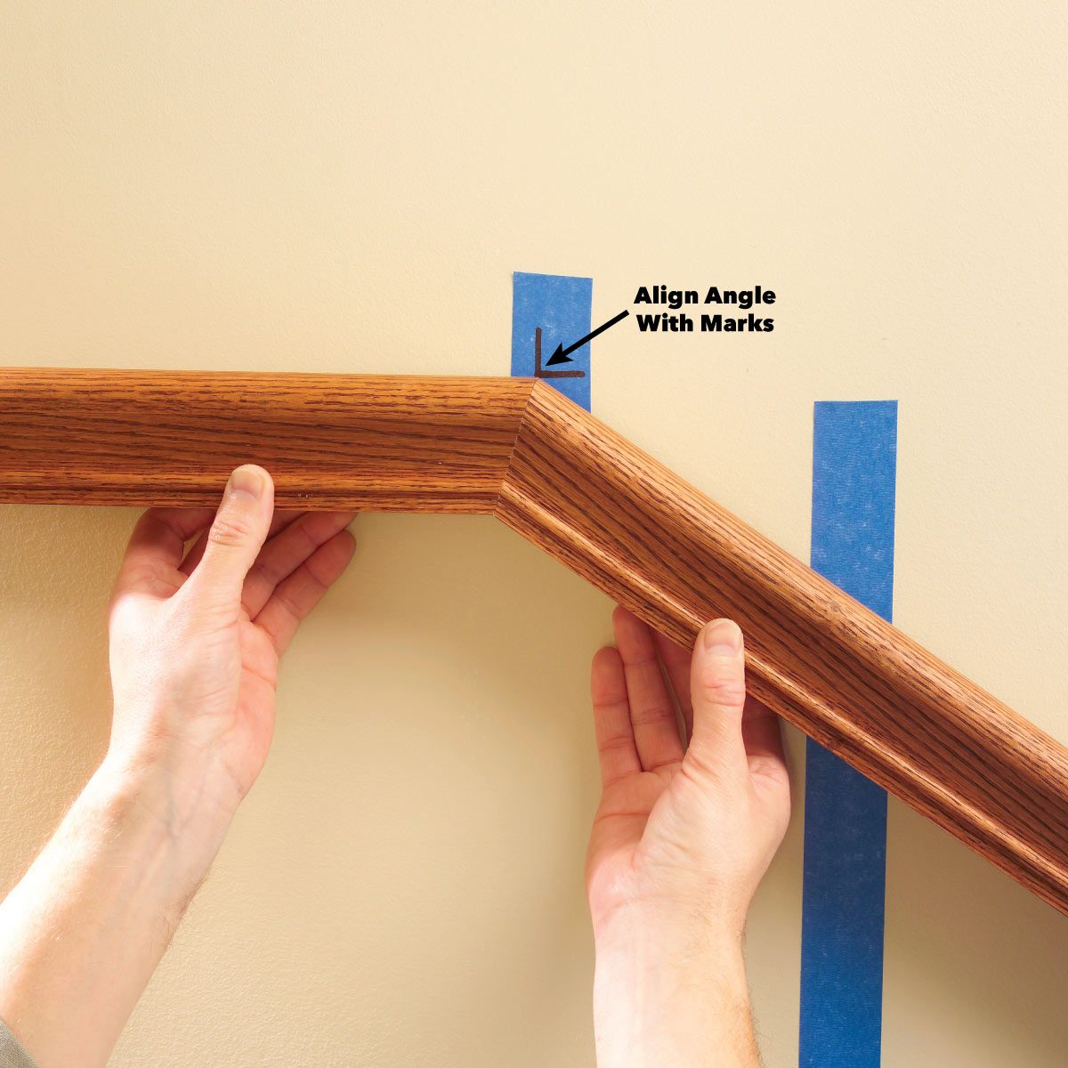 A person aligns a wooden molding piece to blue tape markings on a wall, preparing for installation in a home setting.