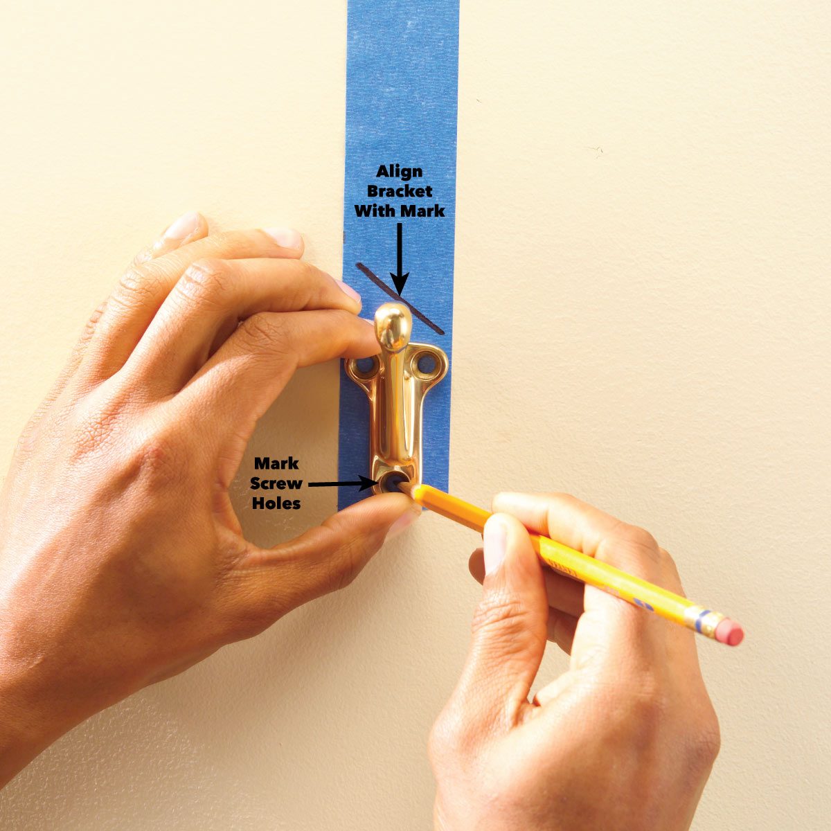 Two hands position a gold wall bracket against blue painter’s tape, while one hand uses a pencil to mark screw holes on a wall.
