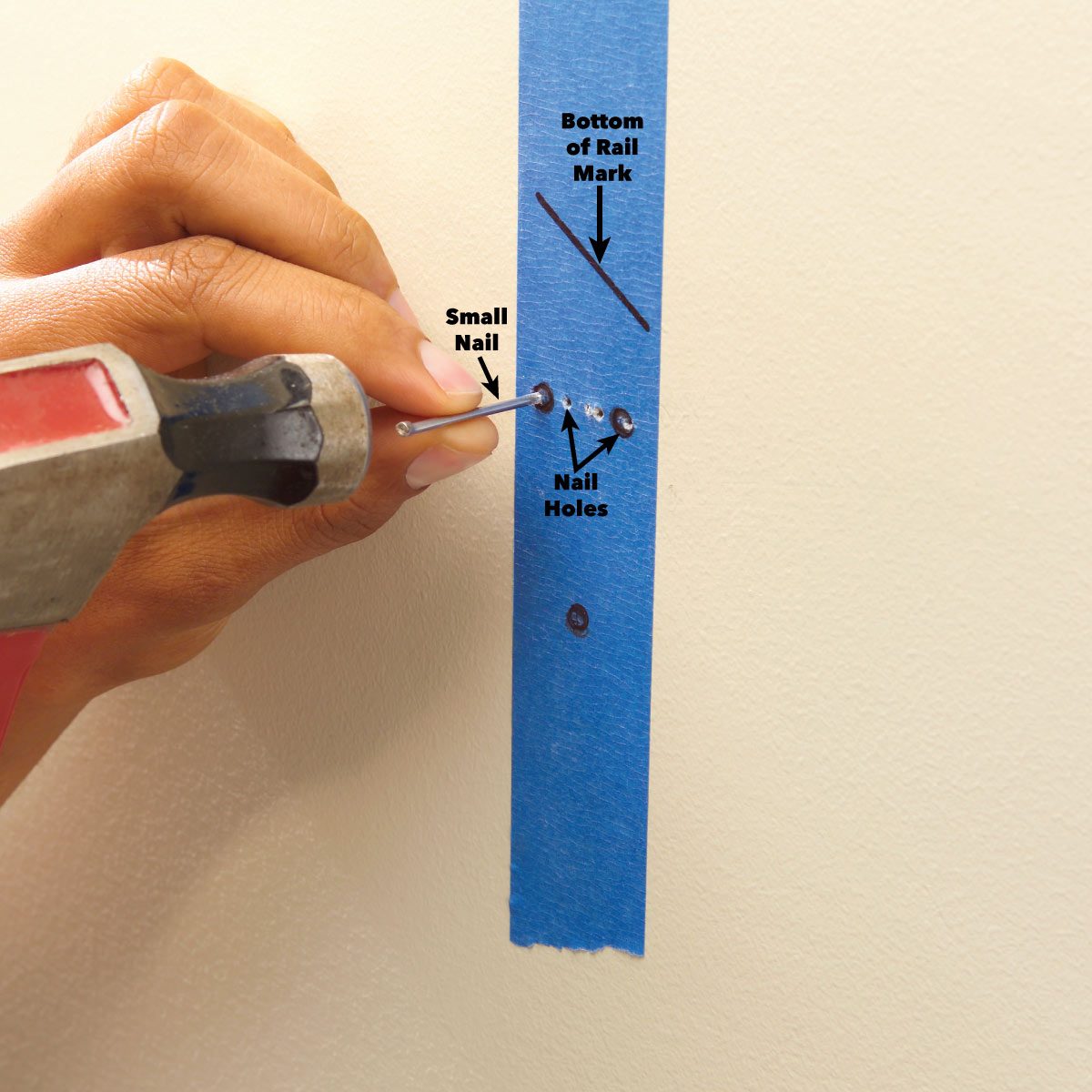 A hand holds a small nail, driving it into wall near blue painter's tape, which marks nail holes and the bottom of a rail.