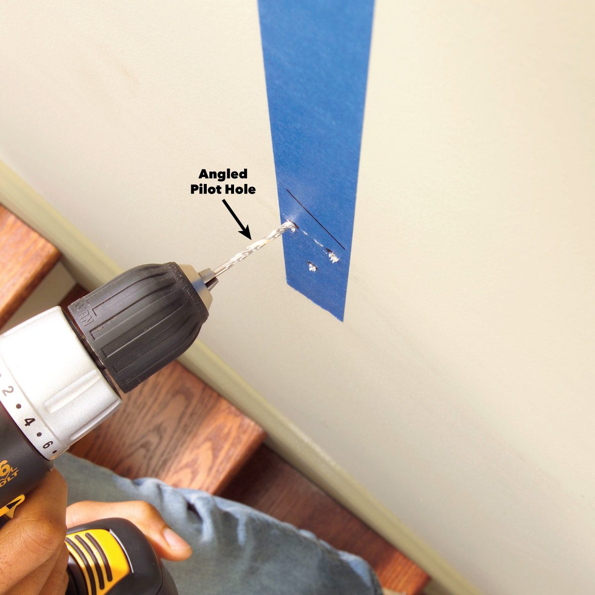 A drill is positioned to create an angled pilot hole in a wall, with blue tape marking the location above wooden stairs.
