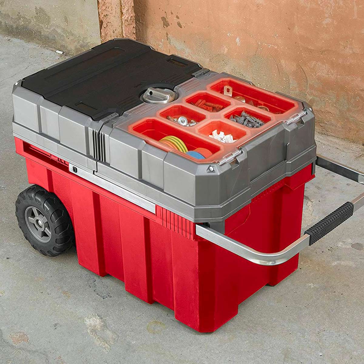 A portable tool storage box with wheels holds various tools and accessories, positioned on a concrete surface against a textured wall.