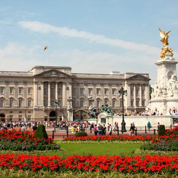 A grand building with intricate architecture stands behind vibrant red flower beds while crowds gather and admire a nearby ornate statue in a sunny outdoor setting.