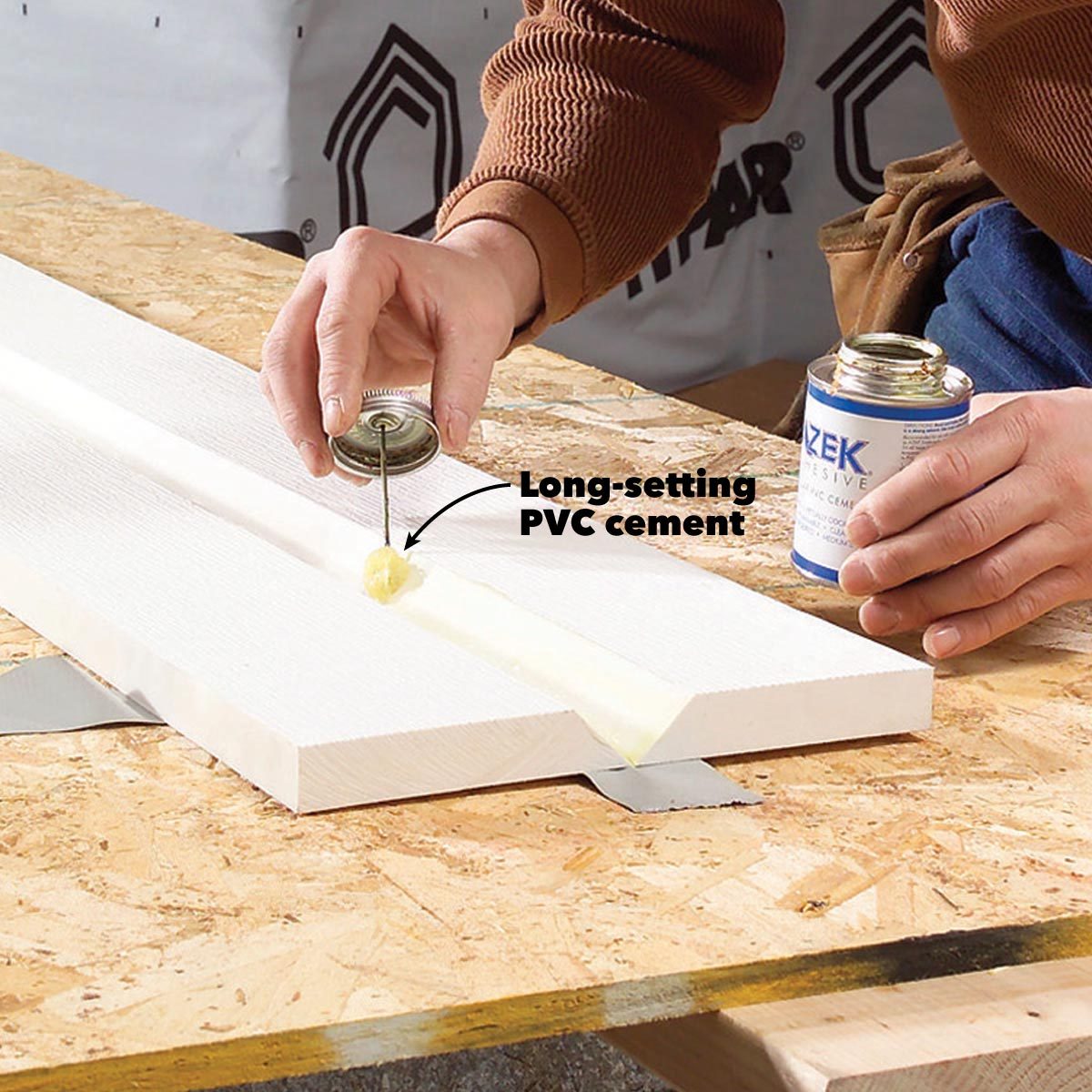 A hand applies long-setting PVC cement to a groove on a white board, while another hand holds a can of adhesive on a workbench.