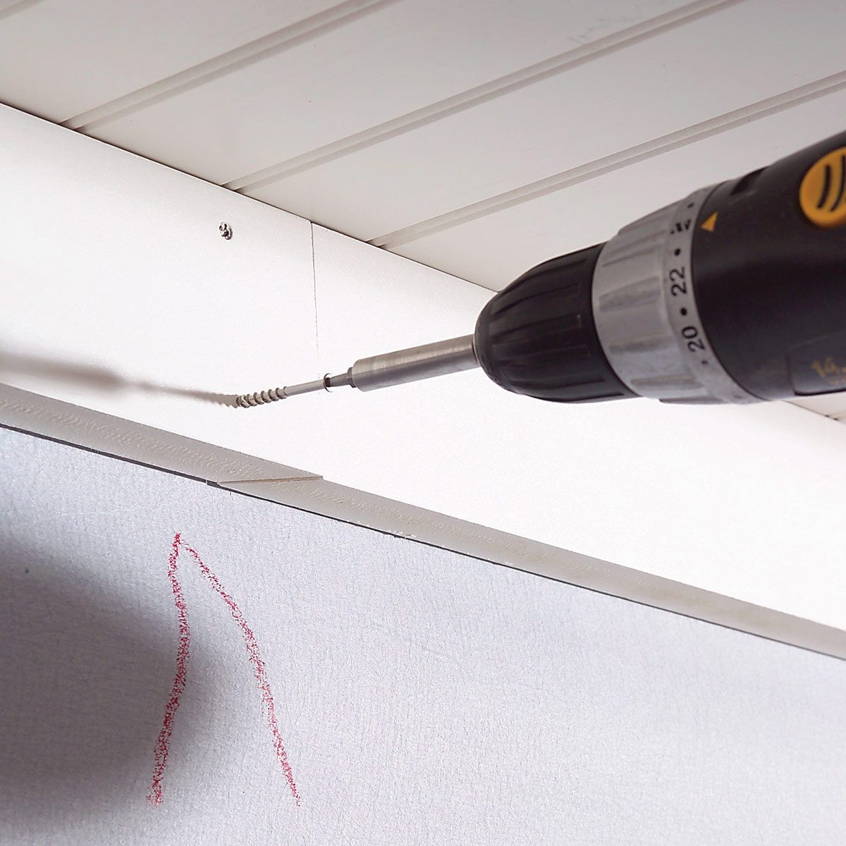 A cordless drill is screwing into a ceiling panel, surrounded by a light-colored ceiling and an unfinished wall with red markings.