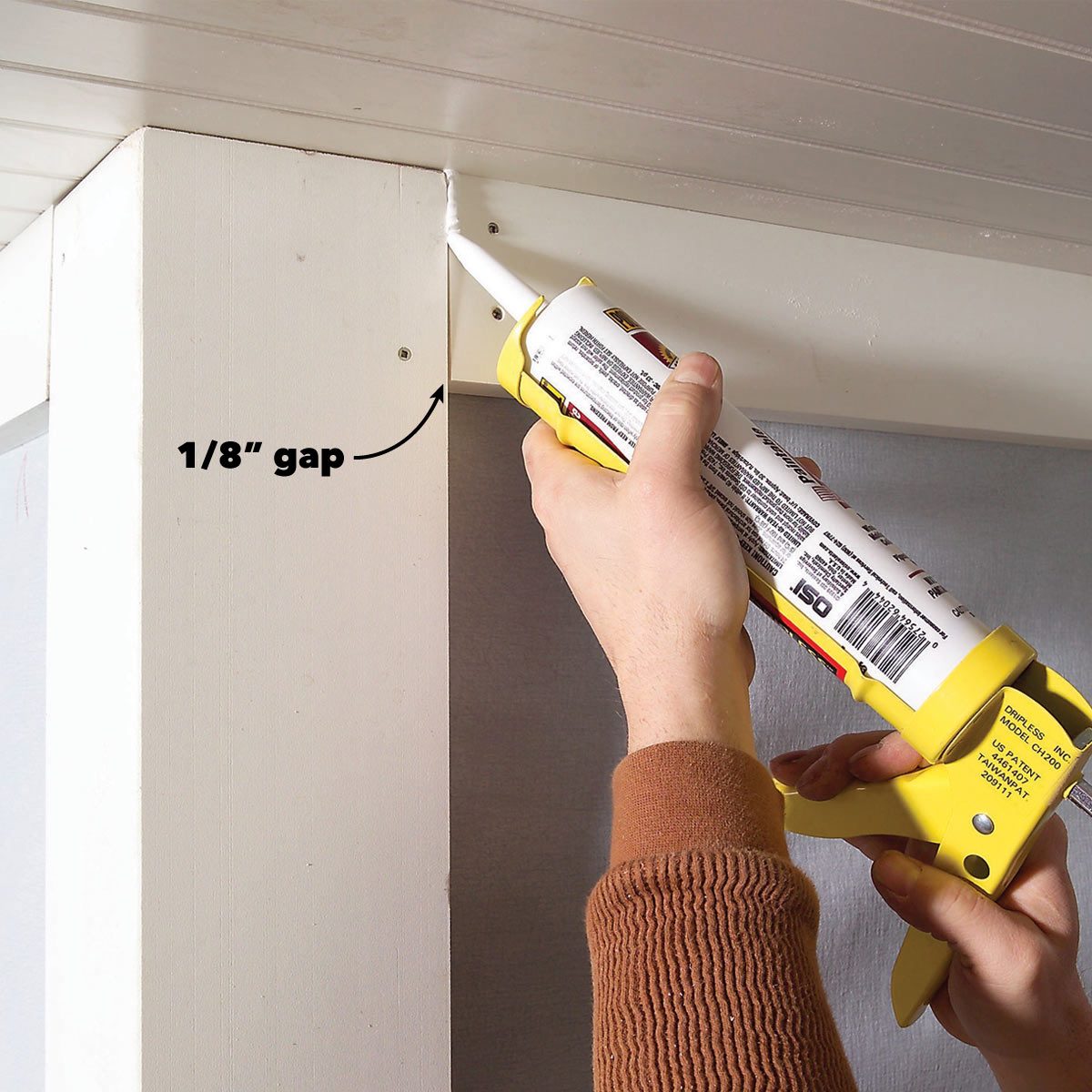 A hand applies caulk from a tube into a small gap at the corner of a wall and ceiling. The surrounding area is unfinished and plain.