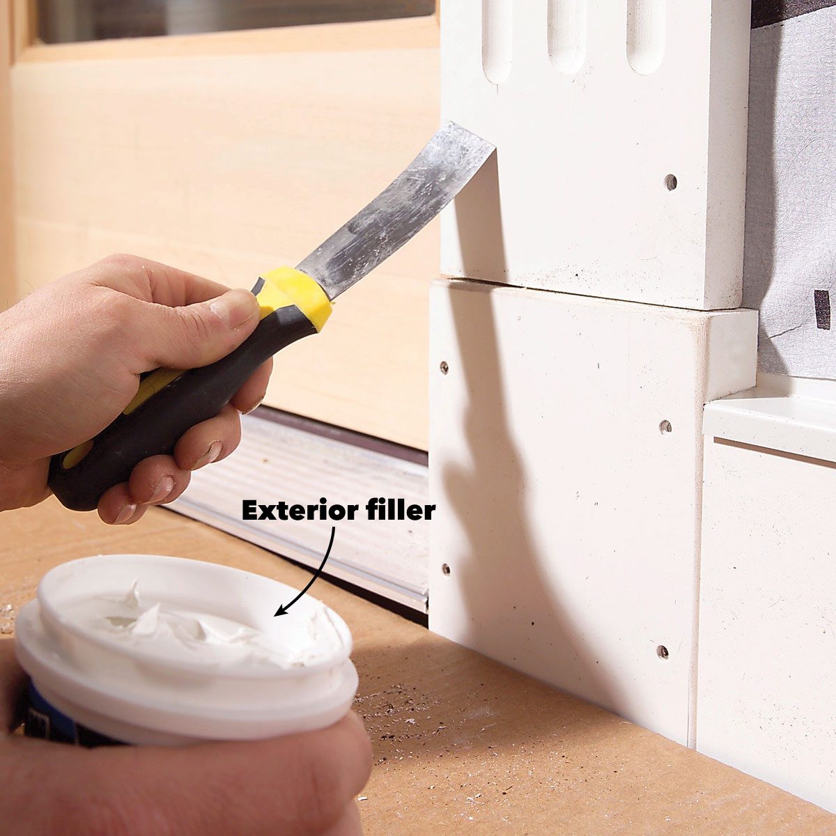 A hand holds a scraper applying exterior filler to a white surface near a door, with a container of filler resting on a wooden surface.