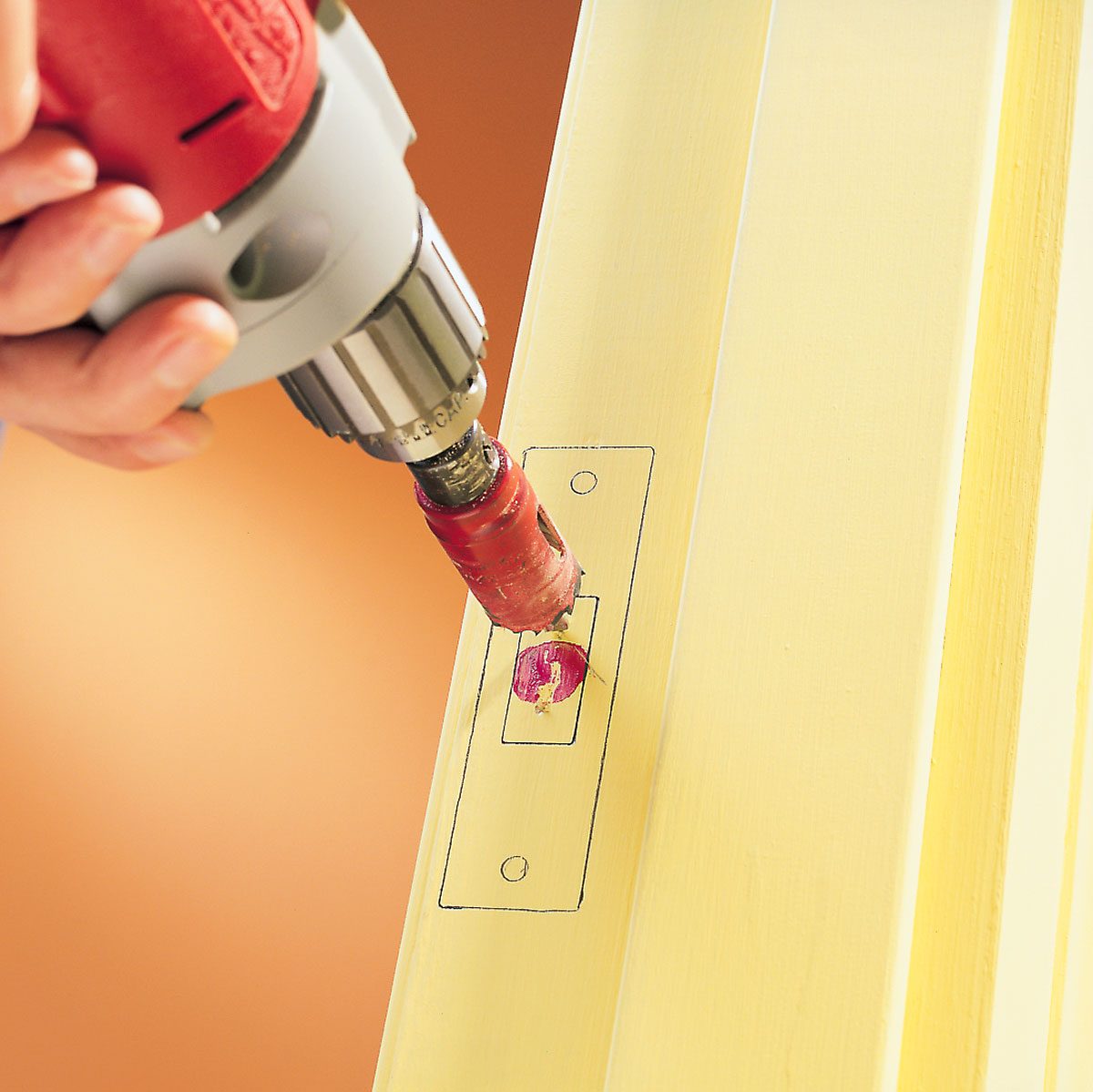 A power drill is being used to create a hole in a yellow wooden surface, outlined with a marked template, against an orange background.