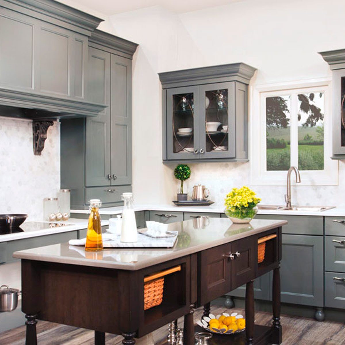 A modern kitchen features a central island displaying bottles and a bowl of lemons, surrounded by stylish cabinetry and a window revealing greenery outside.