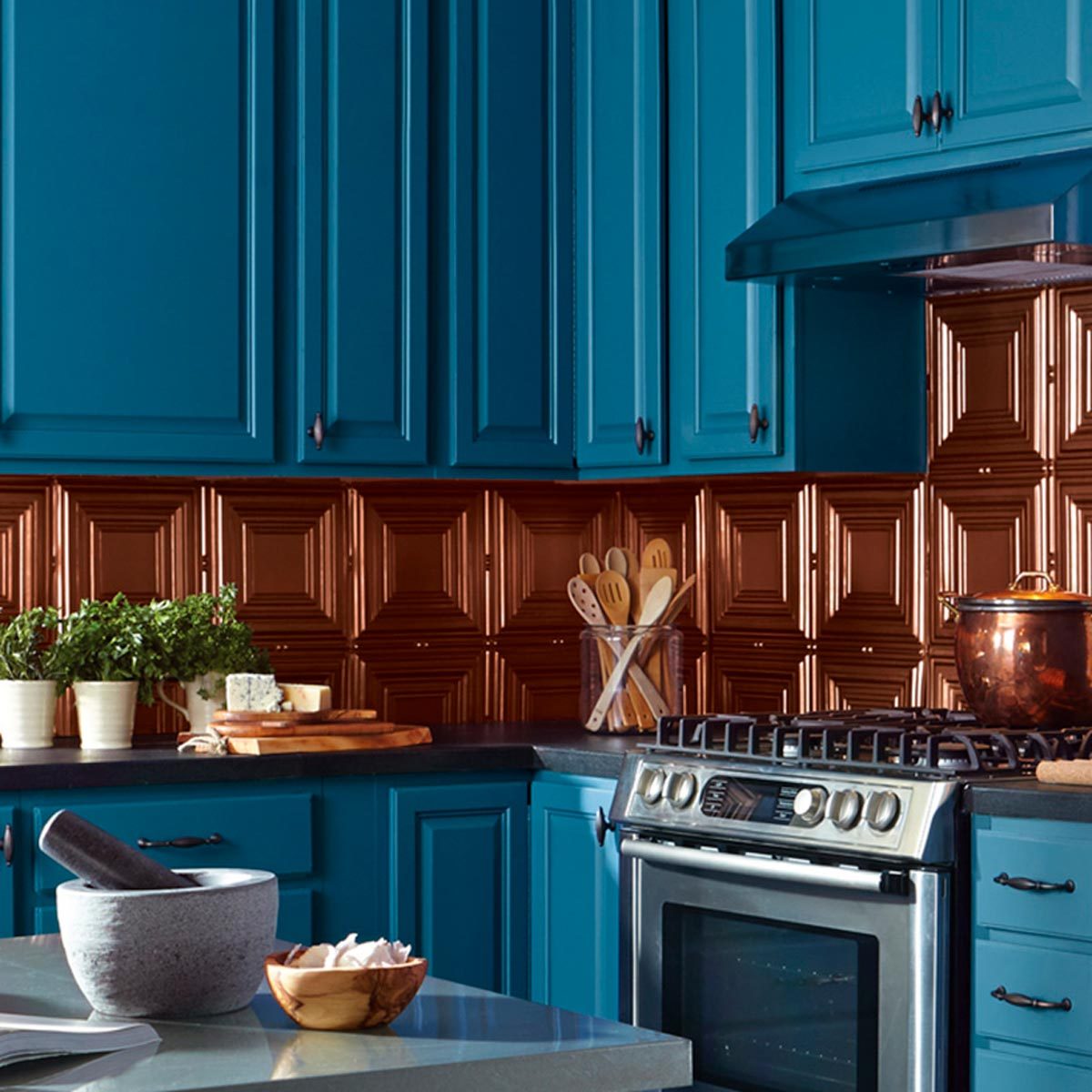 A modern kitchen features blue cabinets above a copper backsplash. A stove sits below, with utensils and fresh herbs on the counter.