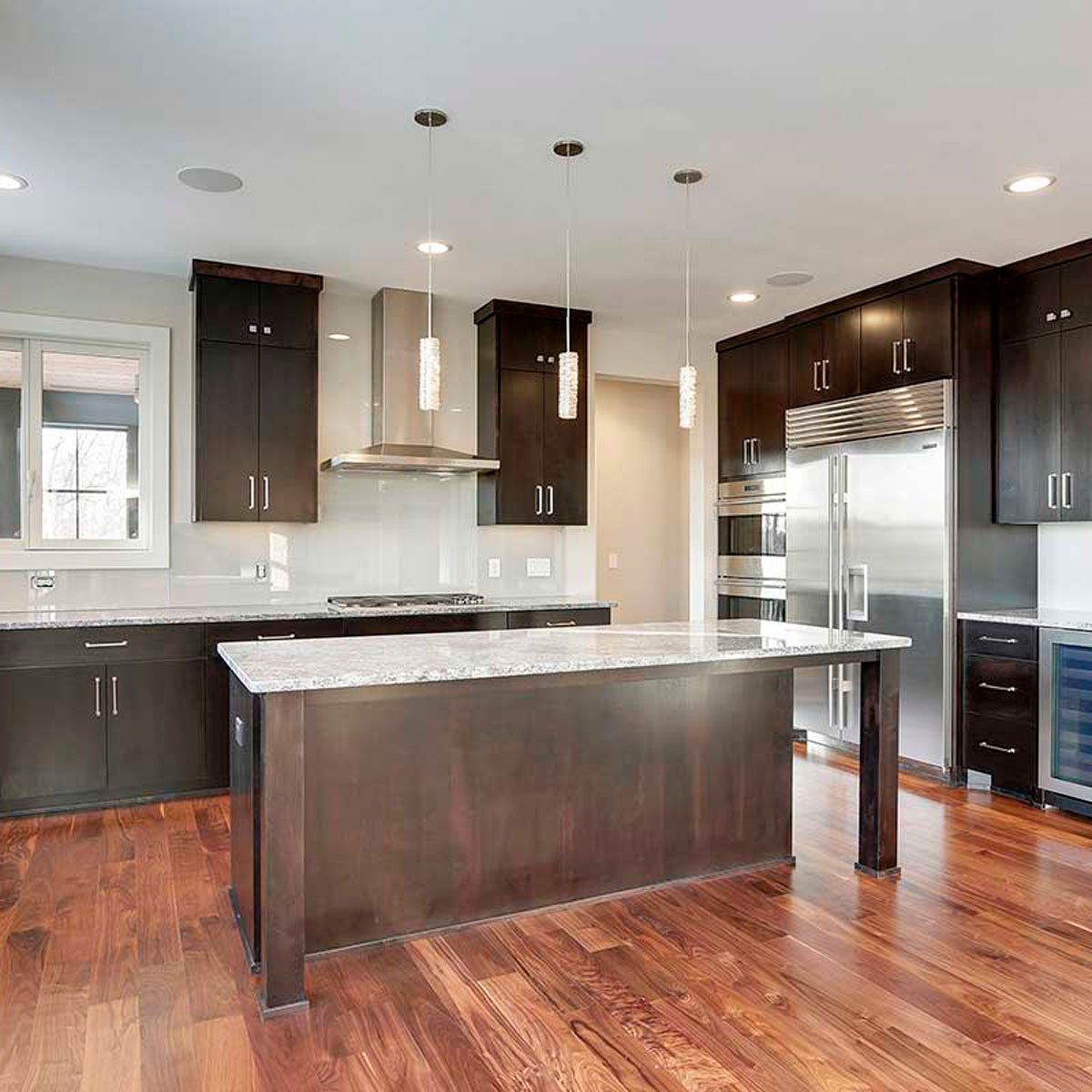 A kitchen features dark cabinetry and a central island with a granite countertop, illuminated by pendant lights, situated on polished wood flooring.