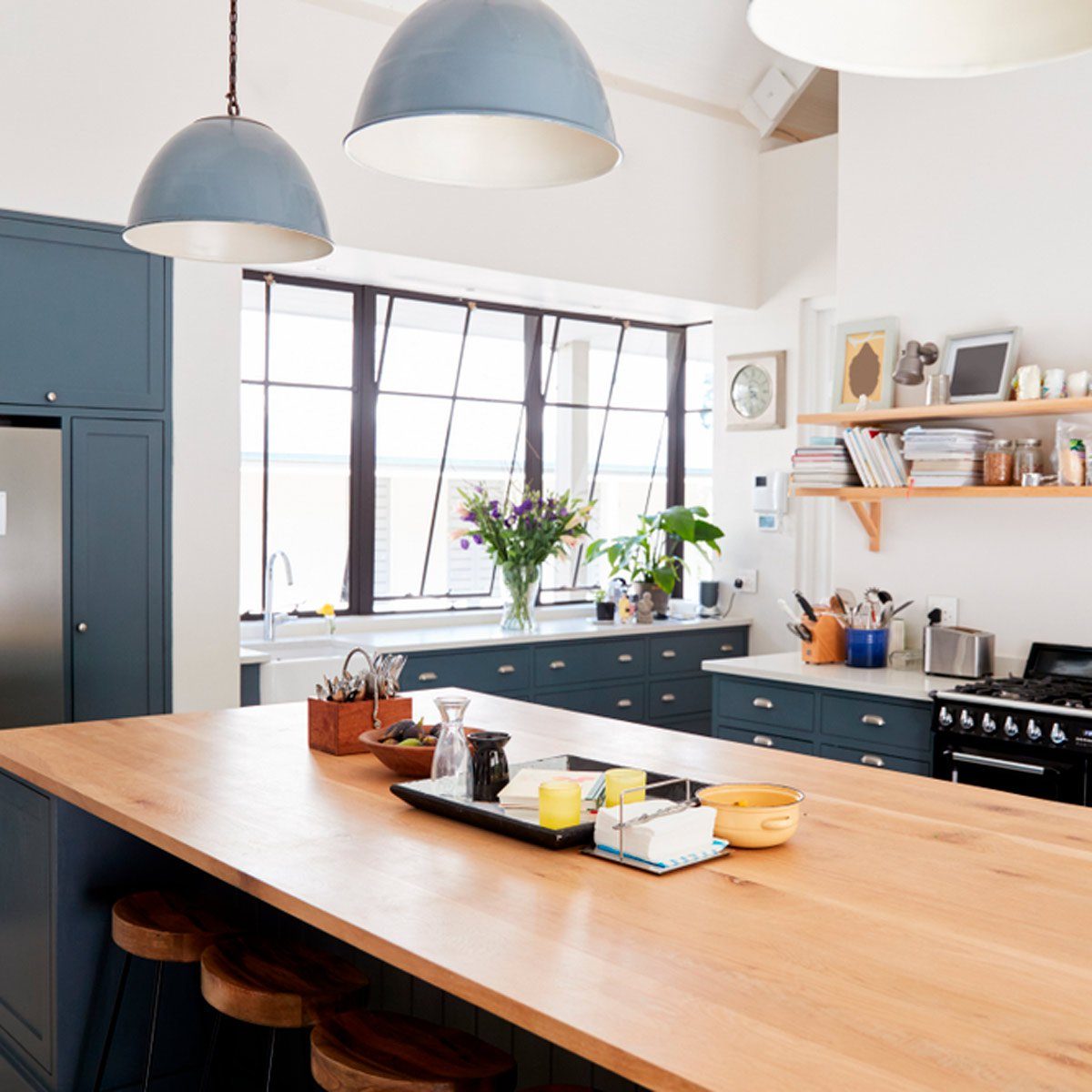 A wooden kitchen island holds a tray with candles and utensils. Light filters through large windows, illuminating blue cabinetry and floral arrangements nearby.