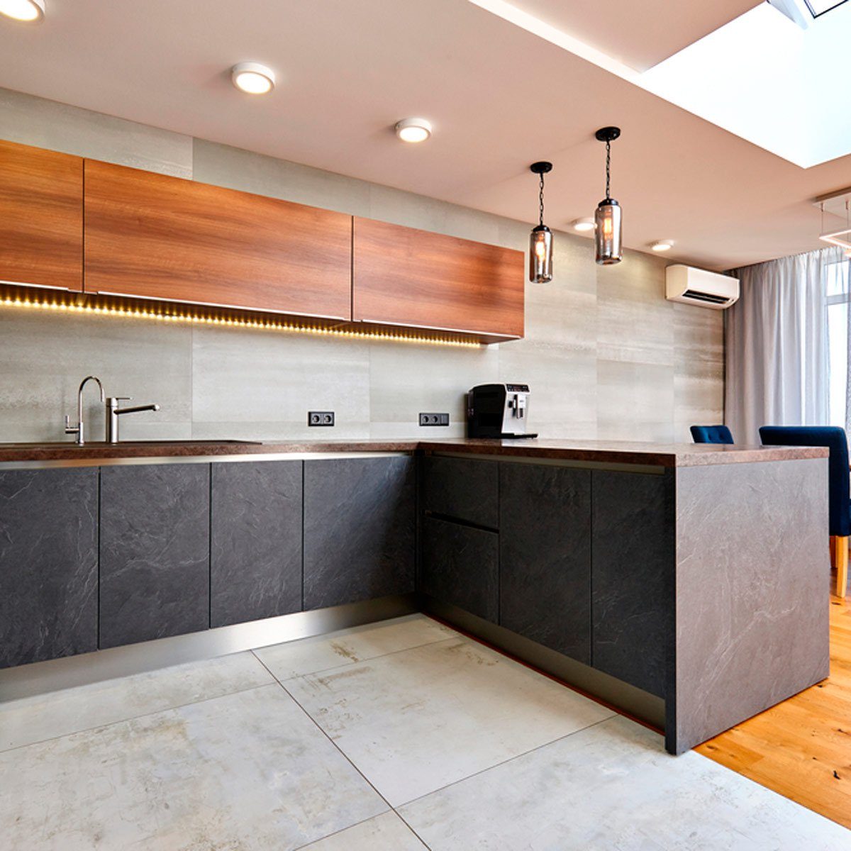 A modern kitchen features a dark stone countertop, wooden cabinets, and industrial-style lighting, with an espresso machine beside a large window and seating area nearby.