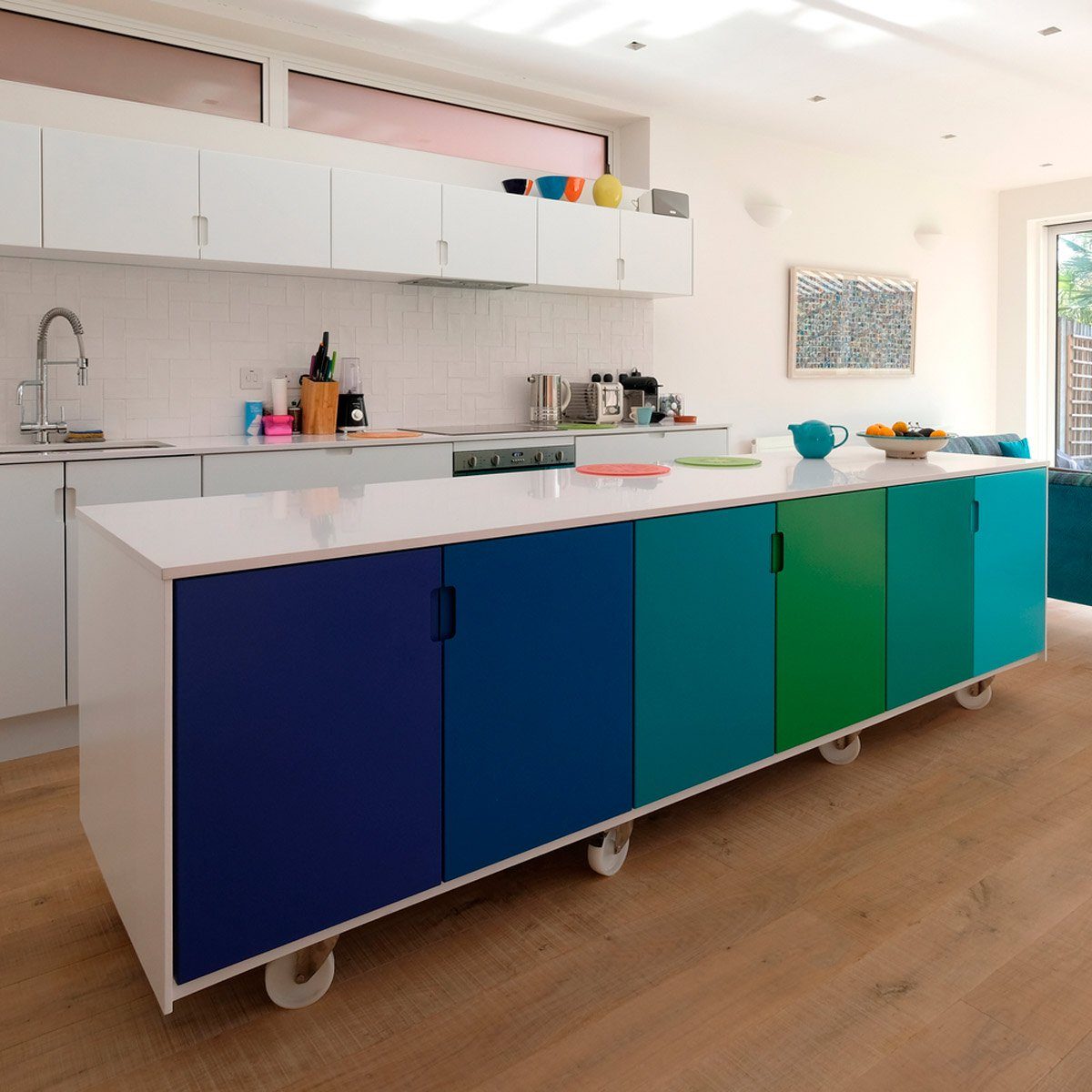 A multi-colored kitchen island with rolling wheels sits in a bright, modern kitchen, surrounded by white cabinetry and a cozy seating area.