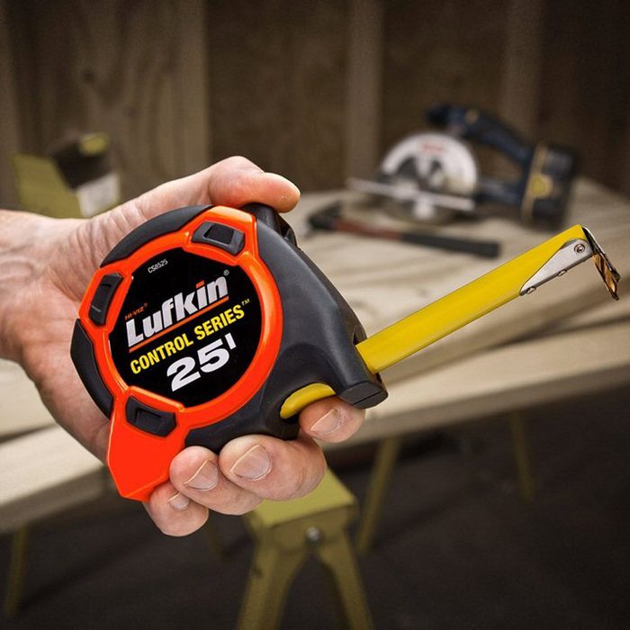 A hand holds a yellow tape measure extending from a black and orange casing, surrounded by tools on a wooden surface in a workshop.