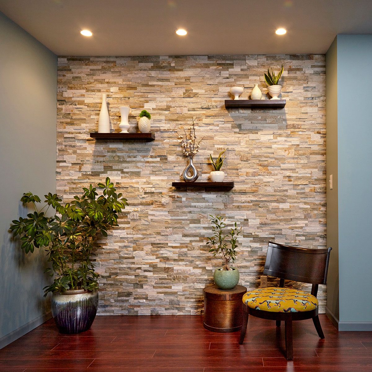 A wooden chair sits on dark wood flooring, surrounded by decorative plants and shelves displaying vases. Soft lighting highlights the textured stone wall.