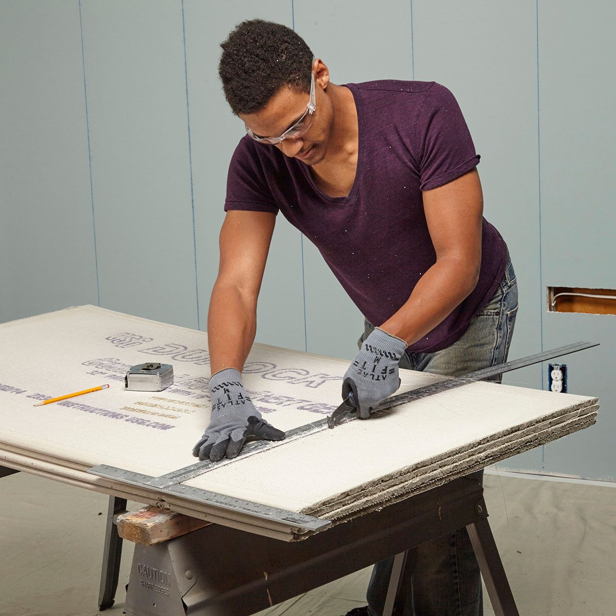 A person cuts a sheet of material on a workbench, using a straightedge and utility knife in a room with light blue walls.