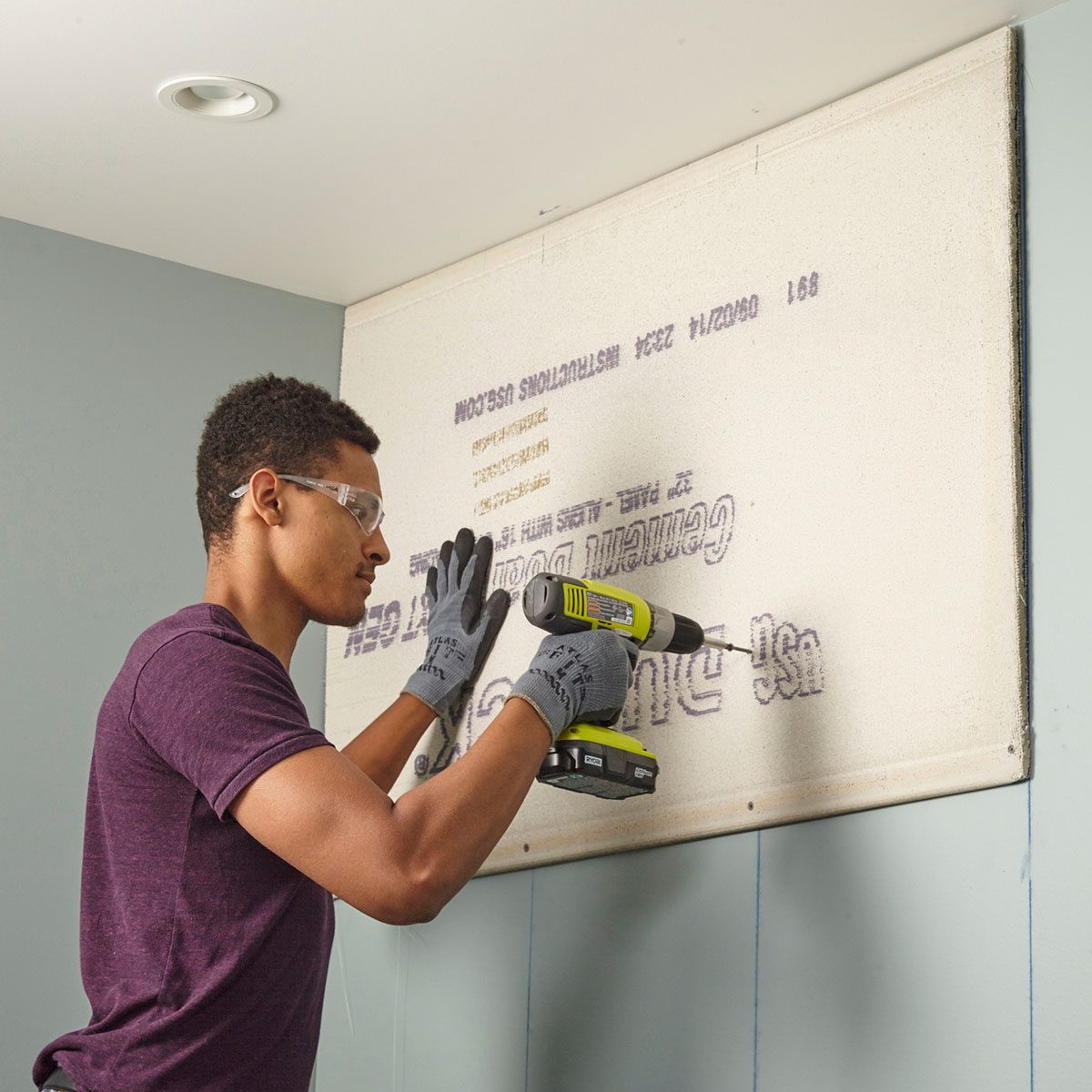 A person uses a power drill to secure a wall panel in a light-colored room with a gray wall. Safety goggles and gloves are worn.