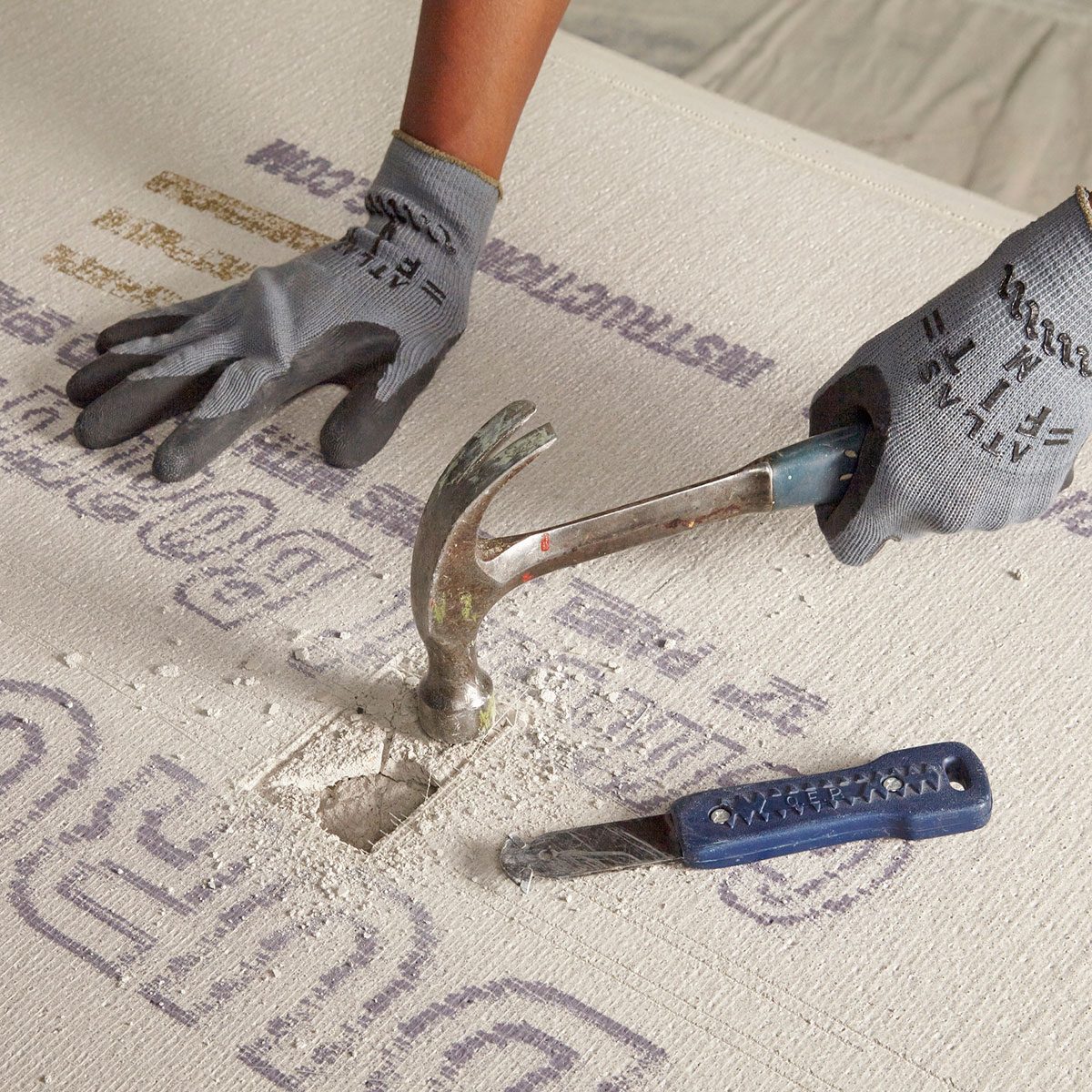 A hand, wearing a glove, hammers a tool into a textured surface, creating dust and a small hole, with another tool resting nearby.