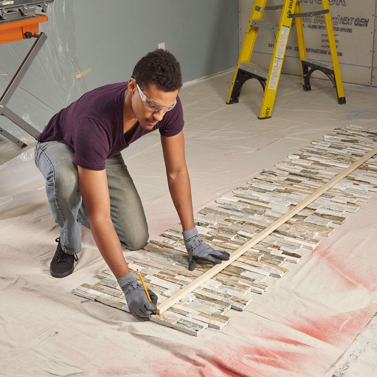 A person kneels, marking measurements on a row of tiles spread on a protective sheet, surrounded by construction tools and a ladder against a wall.