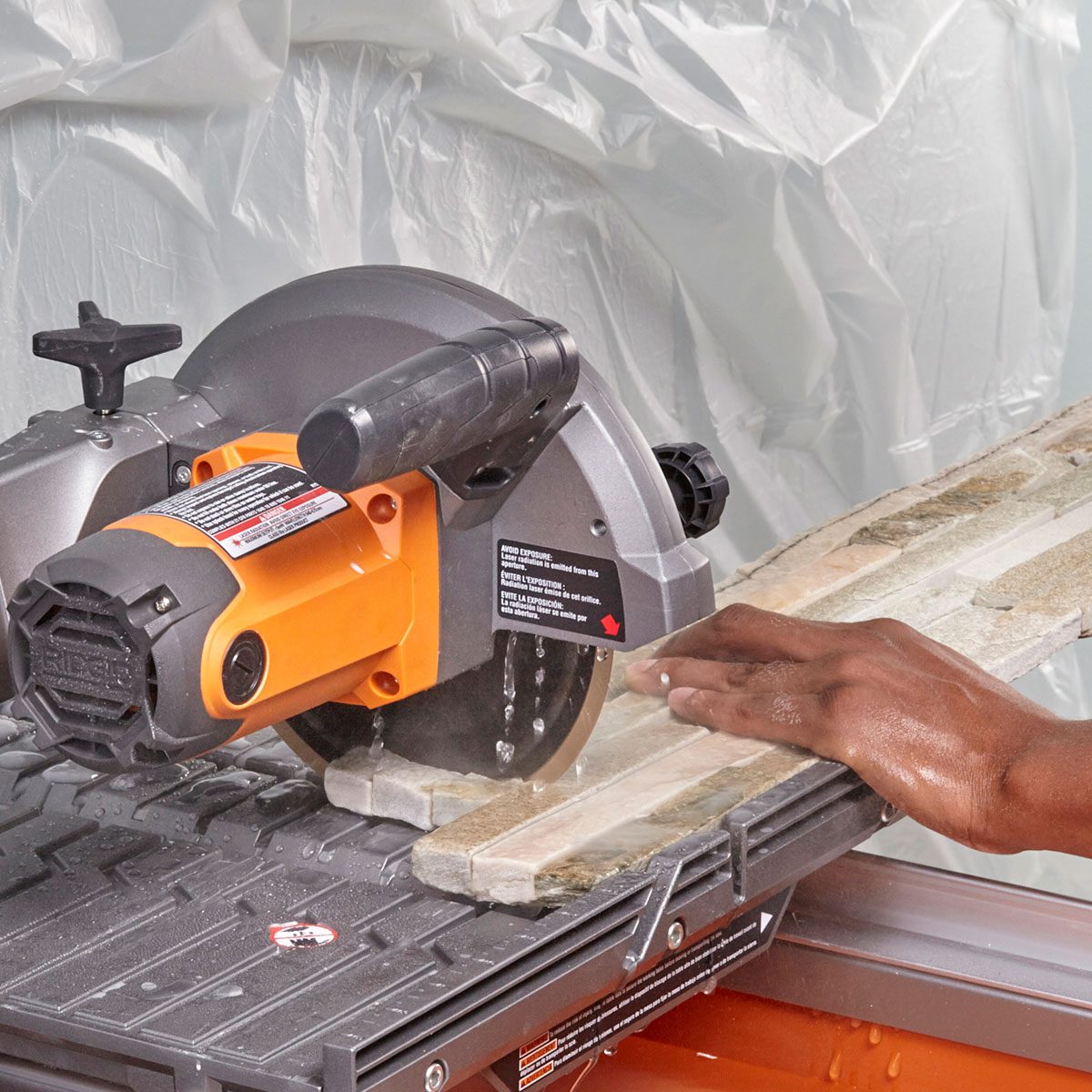 A circular saw cuts through a wooden plank held steady by a person's hand on a work surface, with plastic sheets in the background.