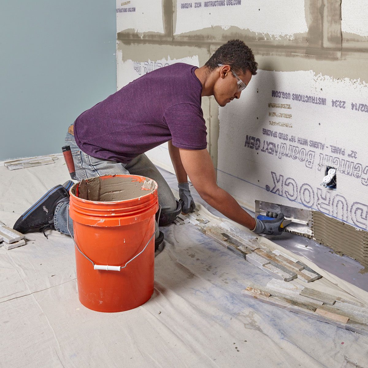 A man kneels, applying adhesive to a wall surface while using a trowel. An orange bucket filled with material sits nearby on a protective drop cloth.