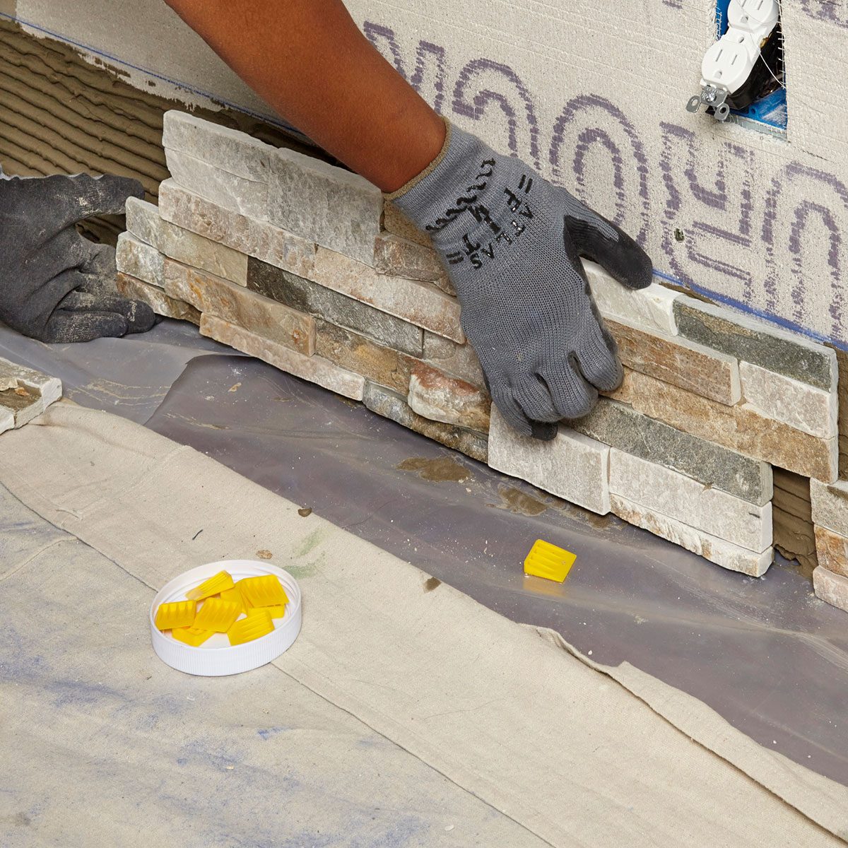 A hand in a glove places stone tiles onto a surface, surrounded by tools and materials, with unfinished electrical outlets visible in the background.