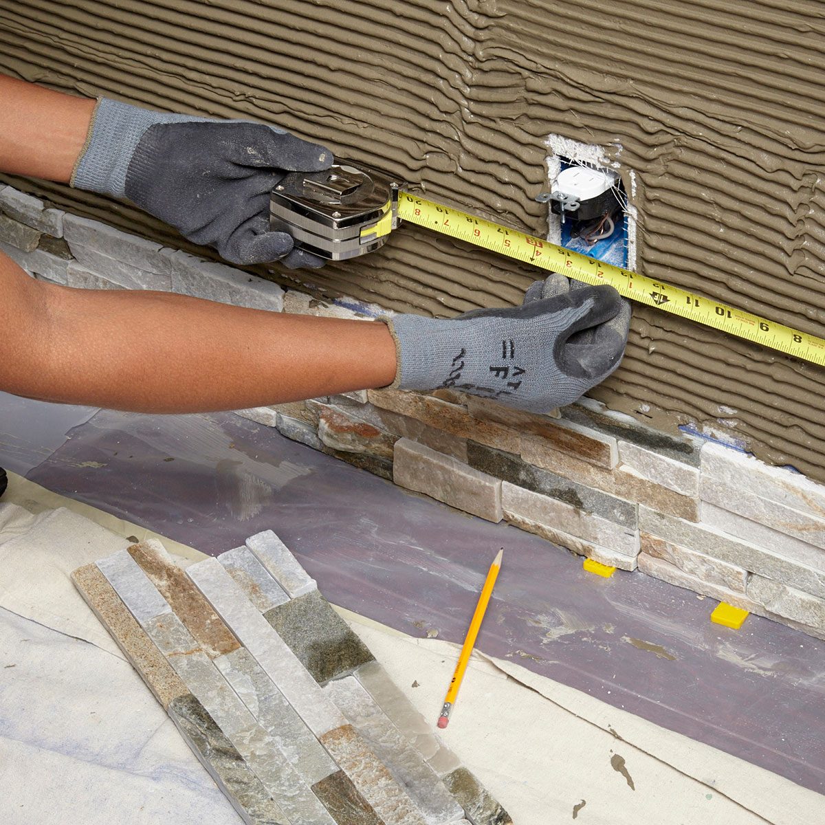 A hand measures a wall with a tape measure while applying stone tiles below. The area is covered with a protective sheet and has a pencil nearby.