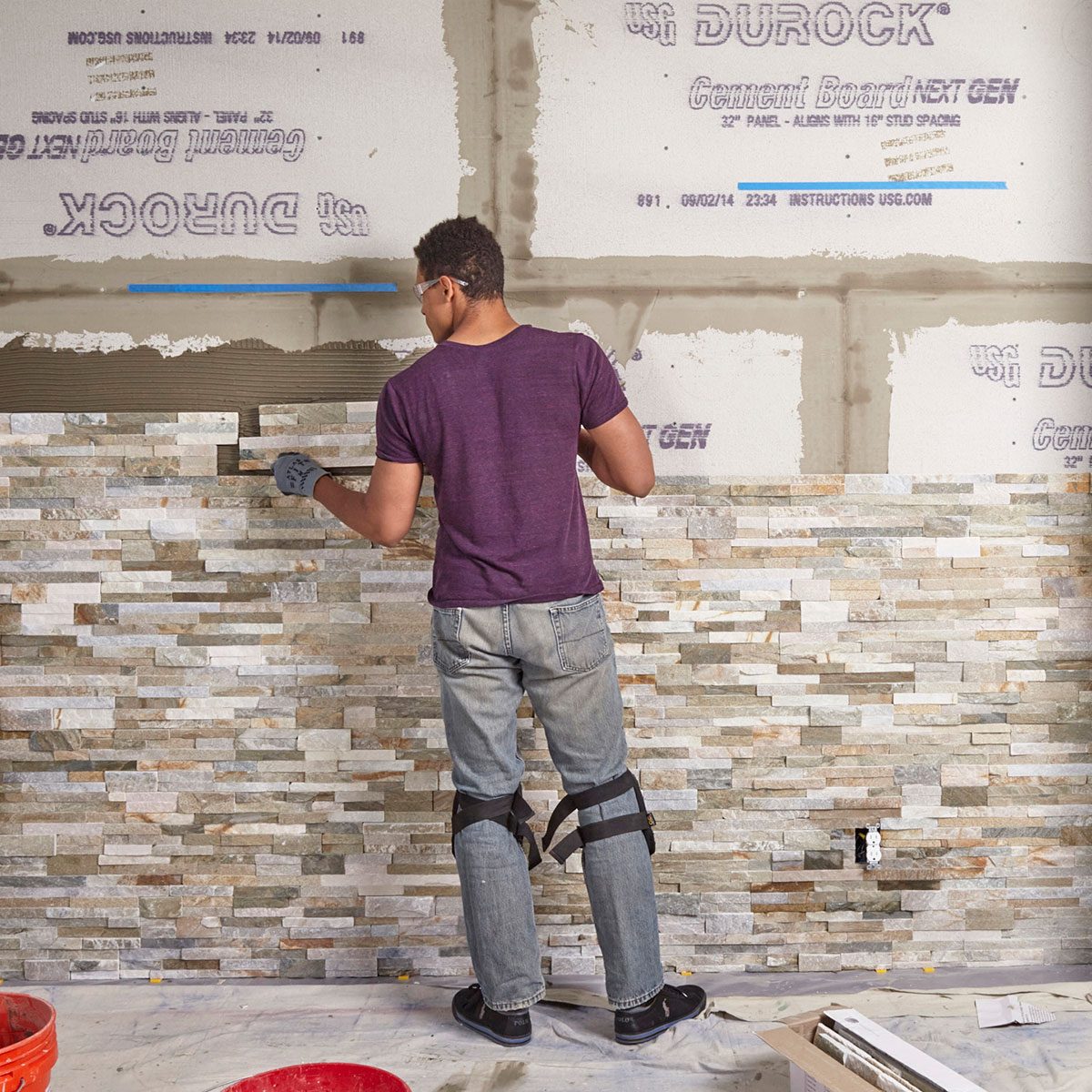 A person applies adhesive while installing stone tiles on a wall in a room with exposed cement board, surrounded by tools and a bucket.
