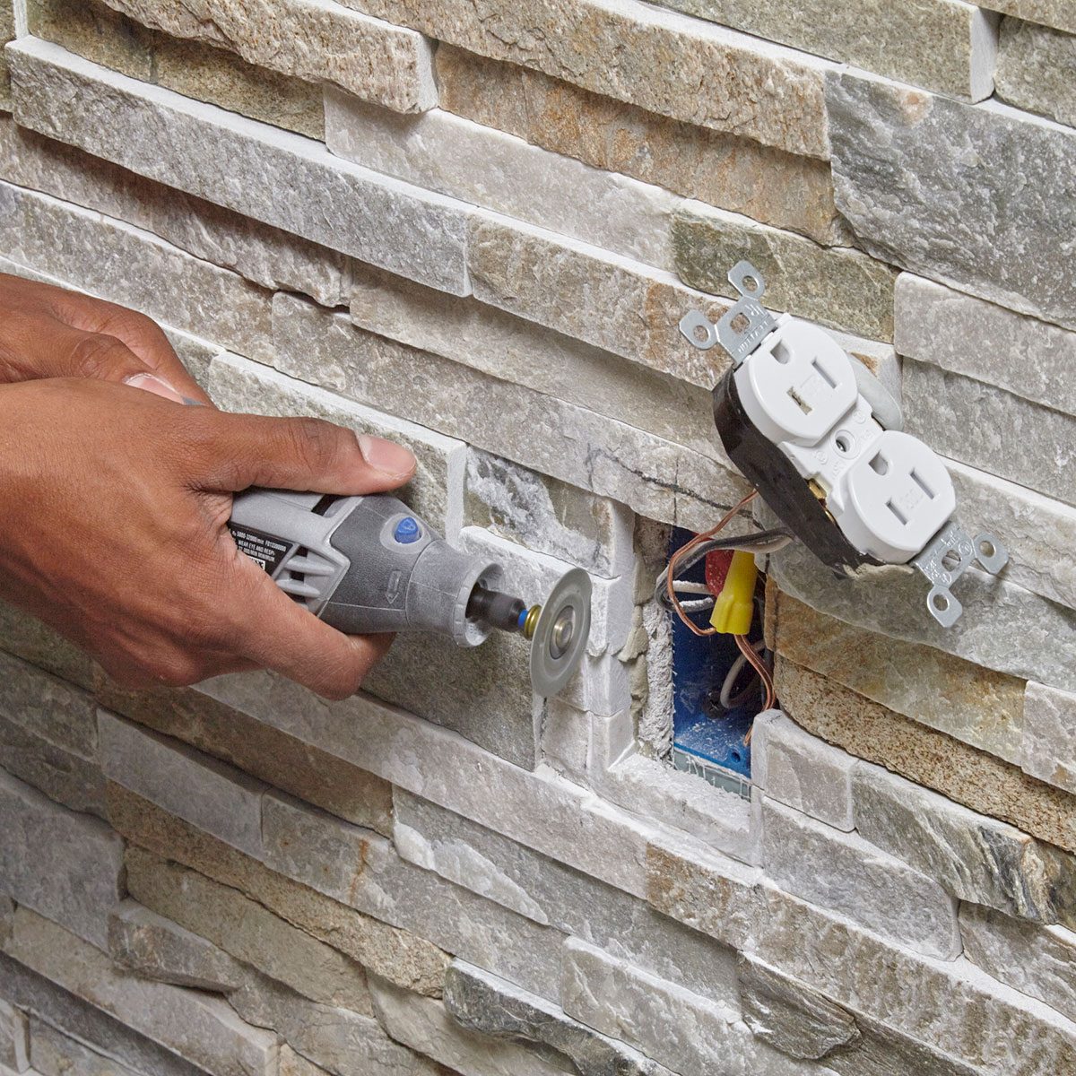 A hand uses a rotary tool to cut into a stone wall, revealing an electrical outlet box amidst textured stone surfaces.