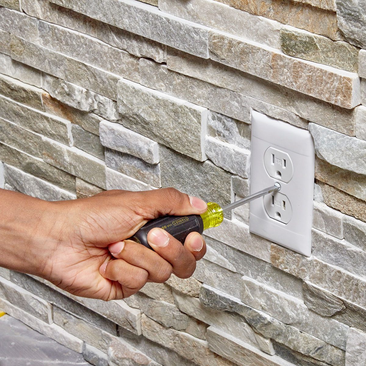 A hand holds a screwdriver, removing a cover plate from an electrical outlet on a textured stone wall.