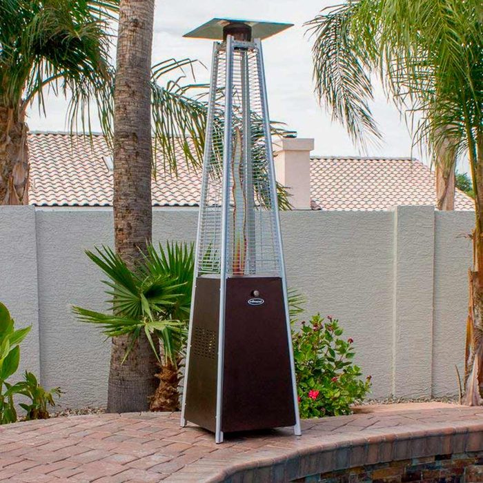 A tall outdoor heater emits warmth, standing beside palm trees and colorful plants in a patio setting, with a stone surface and a gray wall in the background.
