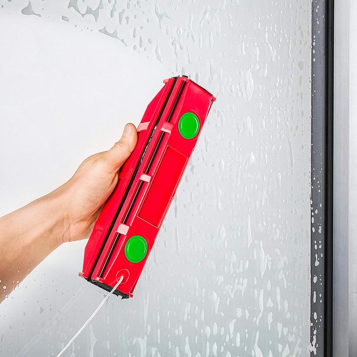 A person's hand holds a red cleaning device, wiping water droplets from a glass surface in a bathroom-like setting.