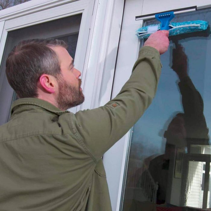 A man cleans a window using a squeegee, focused on removing dirt. The scene is bright, suggesting an outdoor setting, with reflections visible.