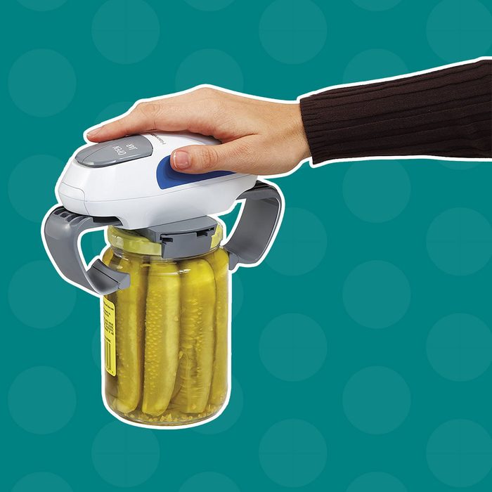 A hand operates a jar opener on a jar of pickles, attempting to twist the lid. The background features a teal pattern.