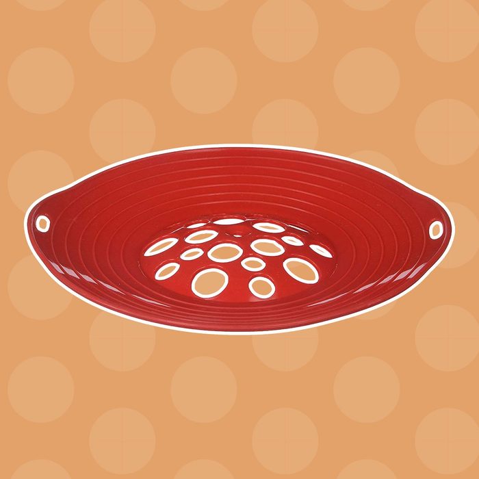 A red colander with circular holes is positioned on a textured orange background with polka dots, designed for draining liquids from food.